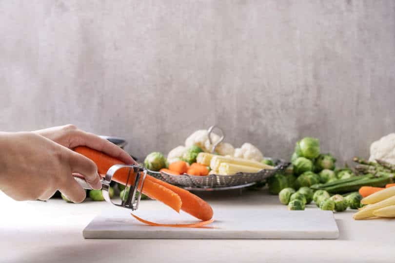 Female hand holding Julienne Peeler served with fresh greens ready to cook: Brussels cabbage, asparagus, baby corn, cauliflower and bamia with metal steamer over a white background.