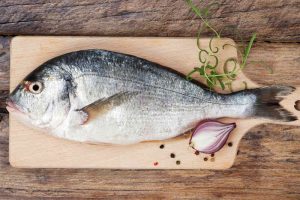 Delicious fresh sea bream fish on wooden kitchen board with onion, rosemary and colorful peppercorns on wooden background.