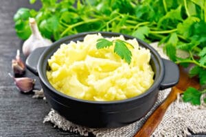 Mashed potatoes in a black pan and a spoon on a burlap napkin, garlic, parsley on dark wooden board background