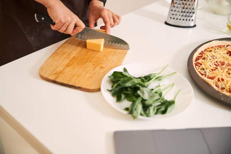 Woman slicing cheese on a wooden cutting board. Pizza and plate of basil by her side