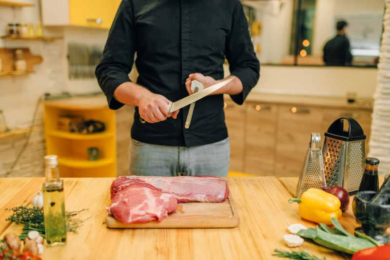 Man with knife prepares to cut raw meat, kitchen interior on background.