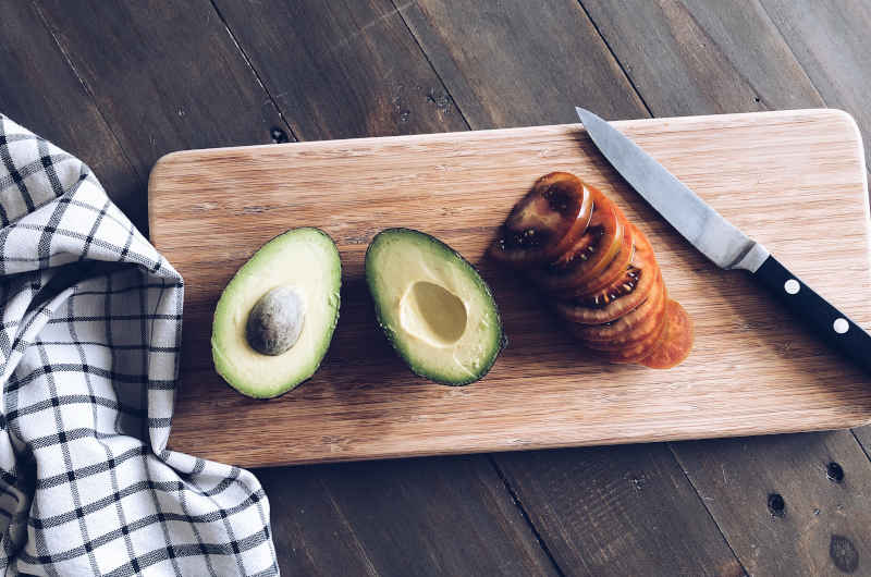 A sliced avocado and sliced tomato with a paring knife on a bamboo cutting board