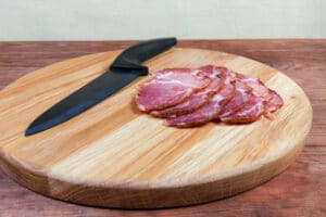 Slices of cured pork neck and black kitchen ceramic knife on the wooden cutting board on rustic table