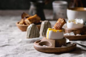 Breakfast with cup of coffee and soft boiled egg, served in wooden egg cup with salt, pepper and toasted bread, jug of cream over linen tablecloth.