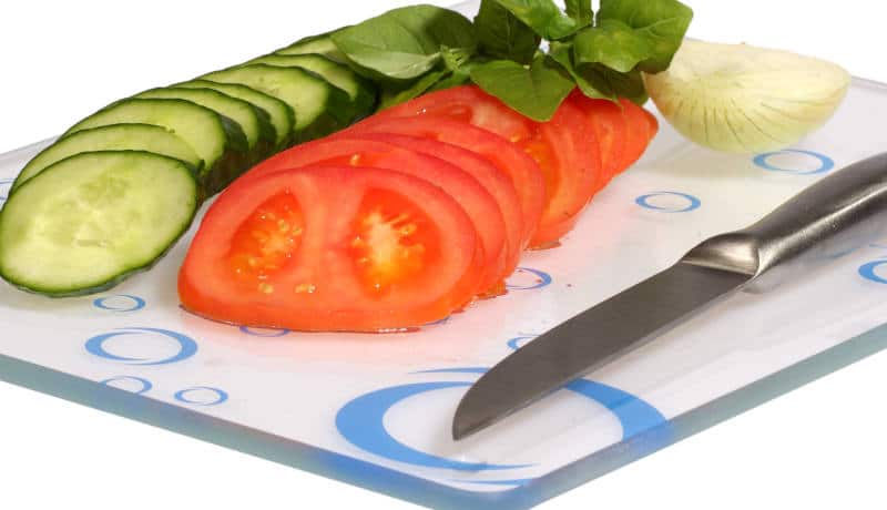 Cucumbers, onions, tomatoes on a glass cutting board over white background