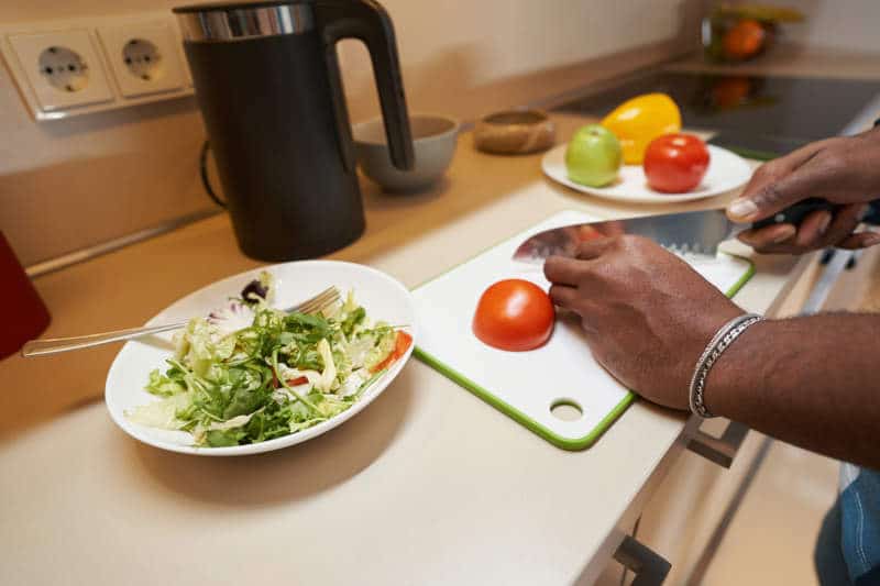 Hand of multiracial person holding knife and cutting red tomato for salad on plastic cutting board